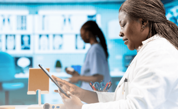 A female medical professional in a white lab coat standing in a high-tech facility, reviewing information on a digital tablet with illuminated medical imaging screens in the background.