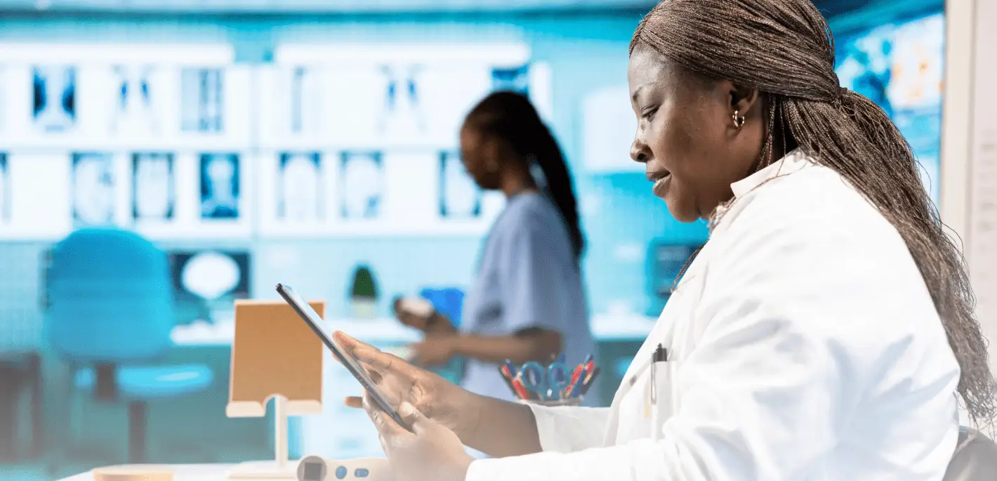 A female medical professional in a white lab coat standing in a high-tech facility, reviewing information on a digital tablet with illuminated medical imaging screens in the background.