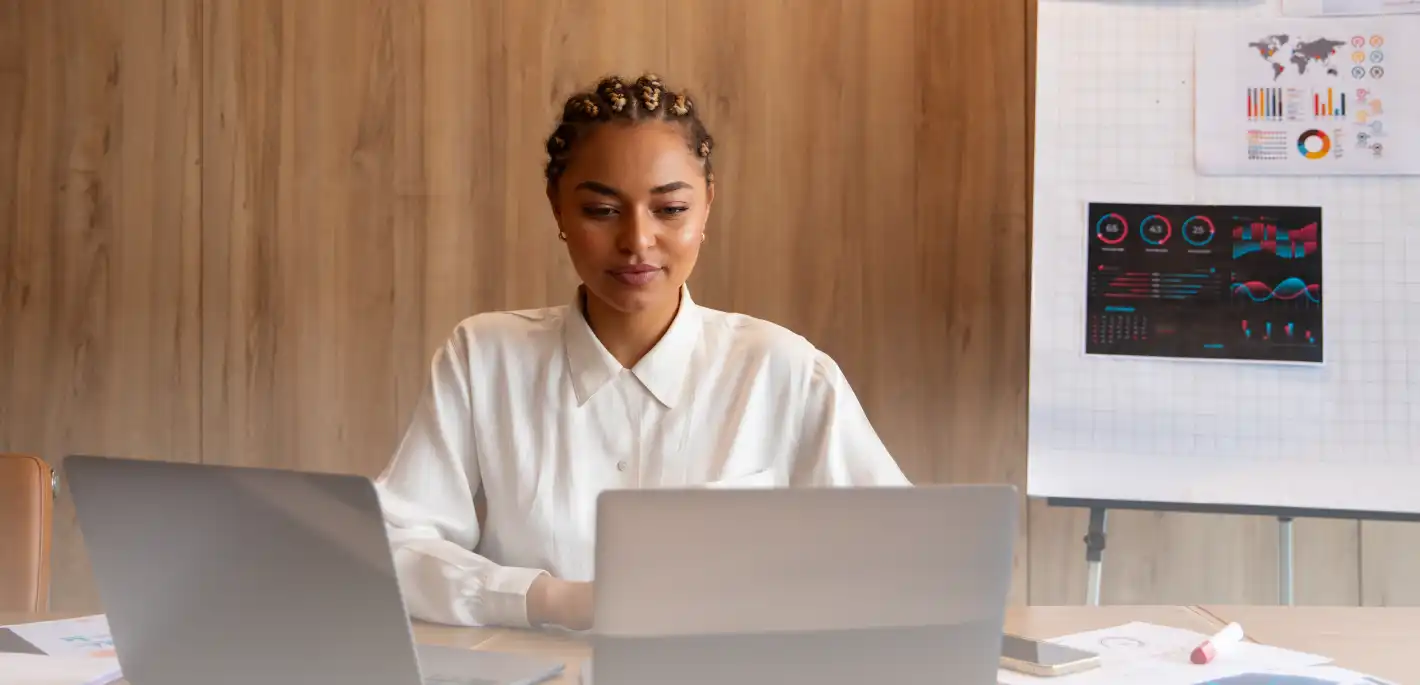 A young woman with braided hair wearing a white blouse, sitting at a desk with two laptops and analyzing data charts displayed on a whiteboard behind her.