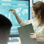 A professional woman points to a "Stock Analyzer" digital screen showing global market charts and a world map while presenting to a group.